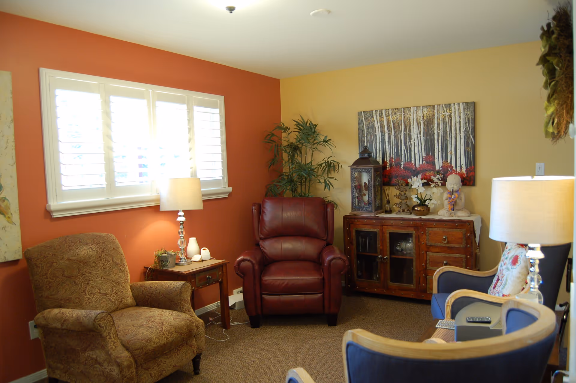 A cozy living room with a red leather armchair, a patterned fabric armchair, and two blue chairs with wooden frames. There is a wooden side table with a lamp and small decorative items next to the patterned armchair. Against the wall is a wooden cabinet with glass doors, decorated with a large lantern, flowers, and a small statue. A painting of birch trees with red foliage hangs above the cabinet. The walls are painted in warm tones of orange and beige, and there is a window with white shutters letting in natural light.
