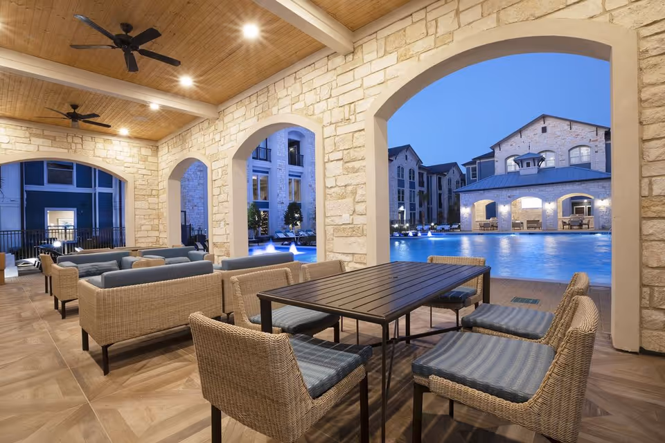 Covered outdoor seating area with wicker chairs and sofas with blue cushions, overlooking a large illuminated swimming pool and stone buildings at dusk.