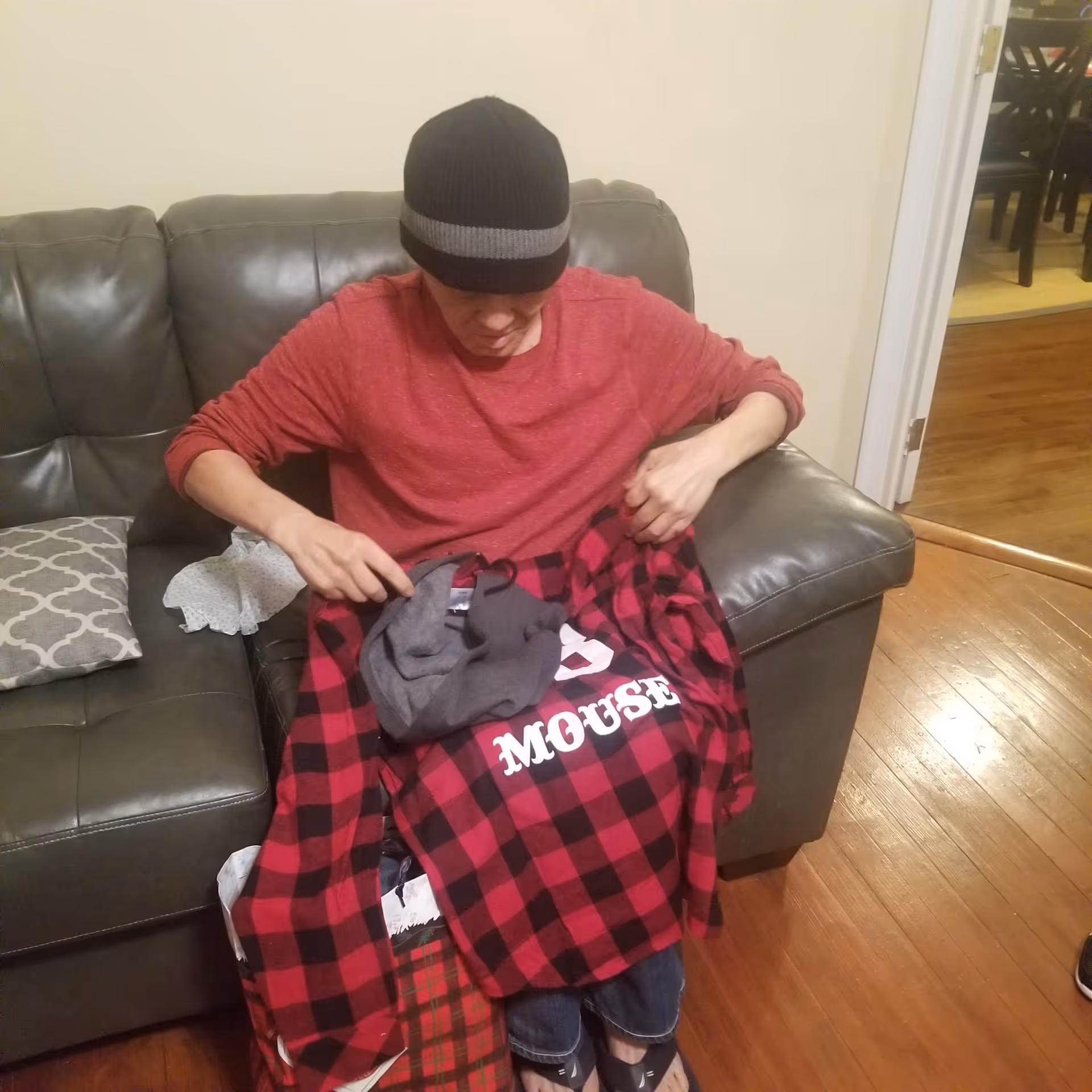 A person wearing a black beanie and red long-sleeve shirt sits on a dark gray leather couch, holding a red and black checkered shirt with the word 'MOUSE' printed on it. There is a gray patterned pillow and a white cloth on the couch beside them. The room has wooden flooring and an open doorway leading to another room with a dining table and chairs.