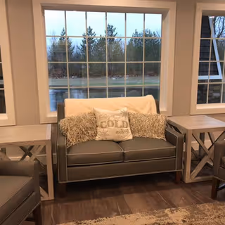 Cozy seating area with a gray loveseat, decorative pillows and matching side tables in front of large windows showing trees outside.