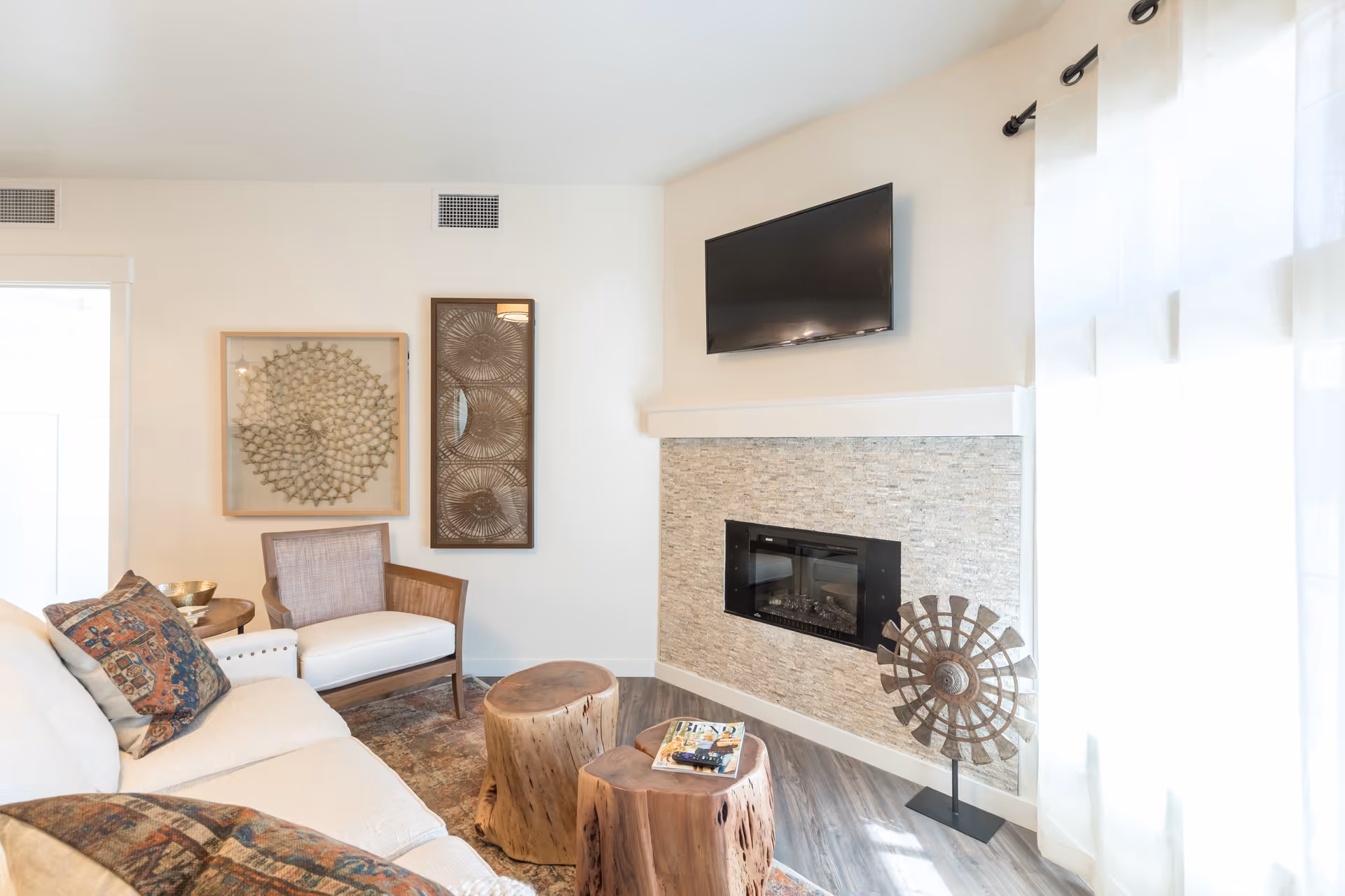 A cozy living room with a beige sofa adorned with patterned cushions, a wooden armchair with white upholstery, two rustic wooden stump coffee tables, a wall-mounted flat-screen TV above a modern fireplace with a stone tile surround, decorative wall art, and sheer white curtains letting in natural light.