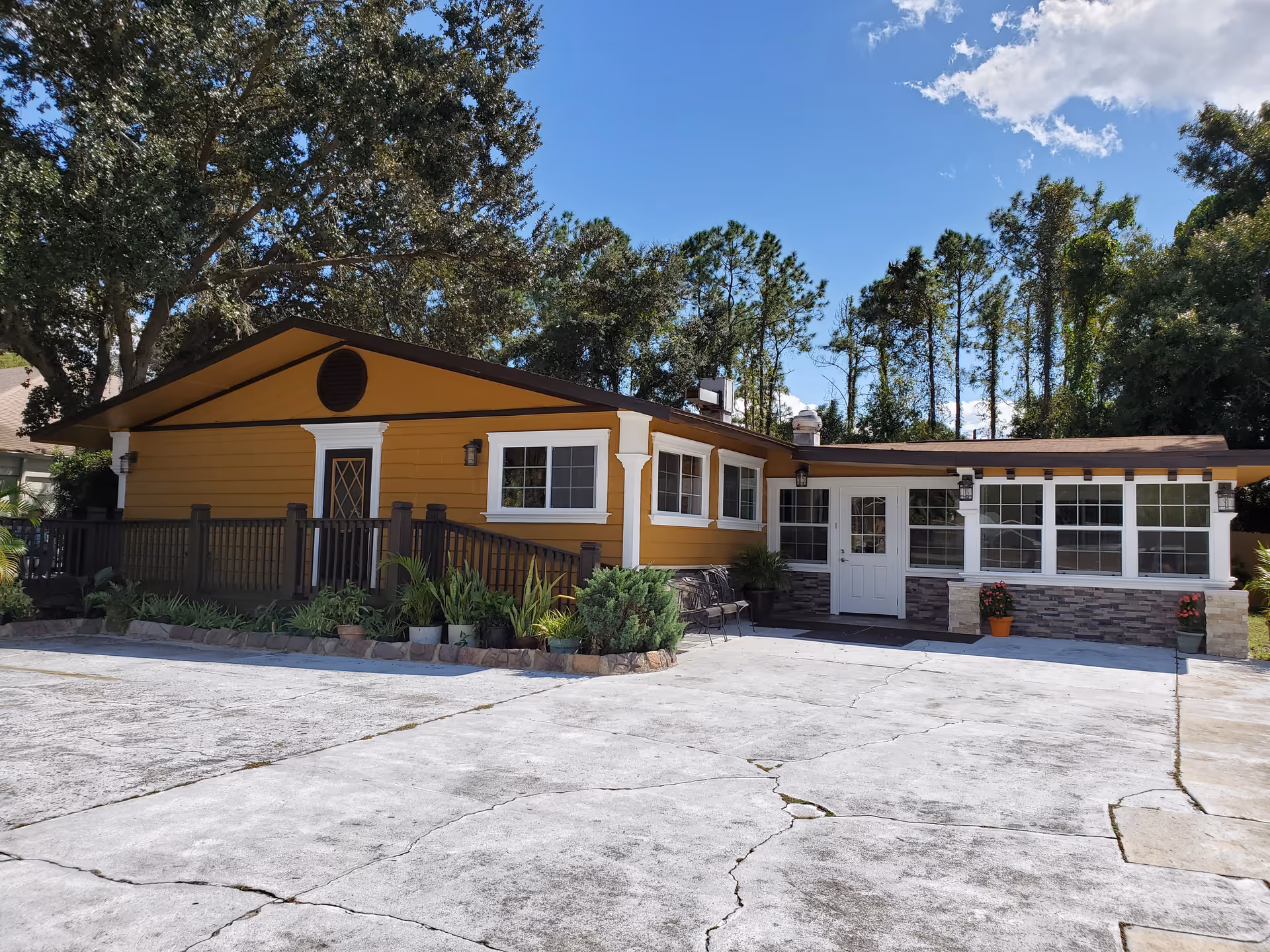 Exterior view of a single-story yellow building with white trim and a brown roof, surrounded by trees and plants. The building has multiple windows, a white door, and a small porch area with a railing and potted plants. The sky is clear and blue with a few clouds.