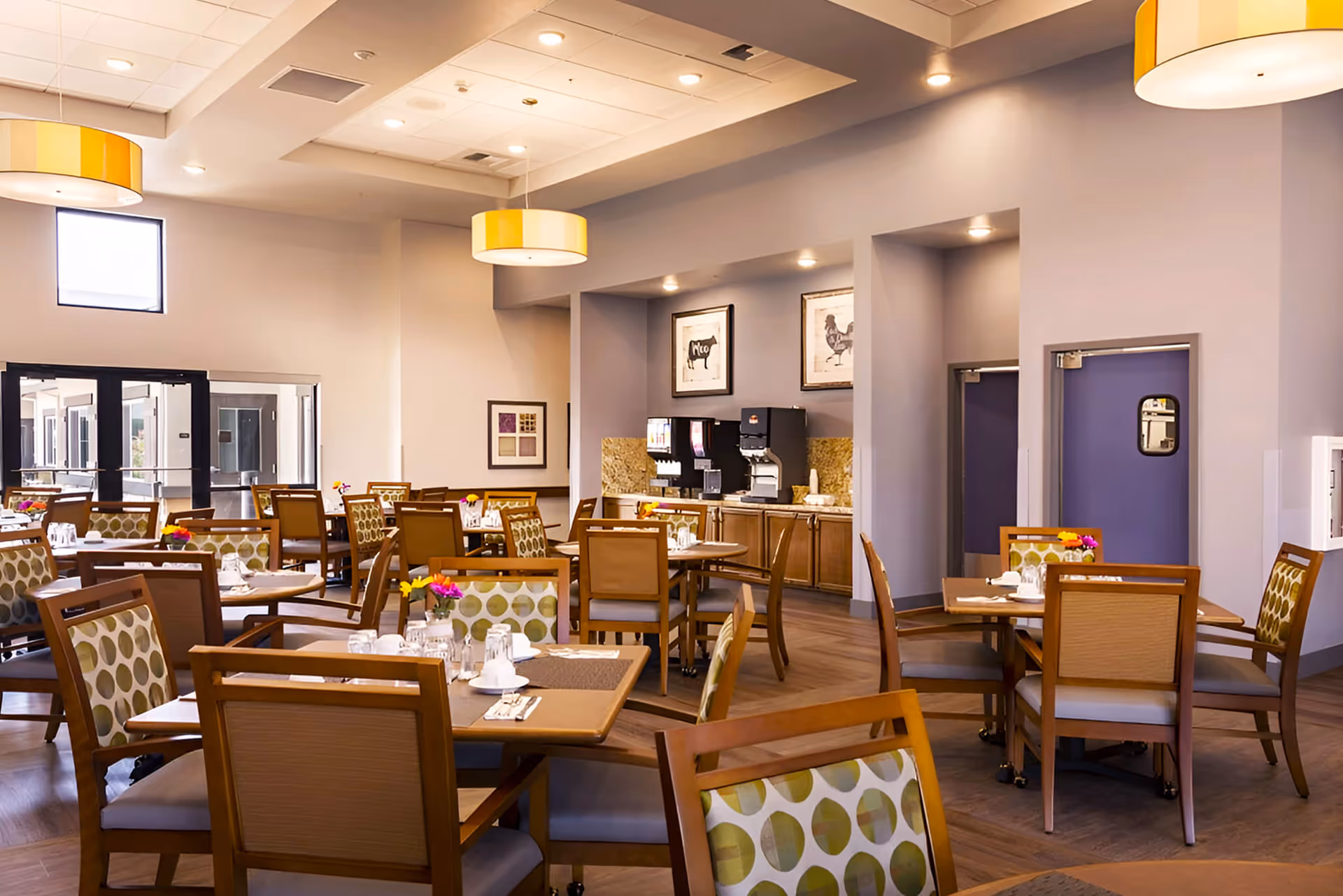 A bright and spacious dining room with multiple wooden tables and chairs, each table set with glasses, napkins, and small flower arrangements. The room features modern pendant lights, light-colored walls, and a beverage station with coffee machines against one wall. Large windows and glass doors allow natural light to enter the space.