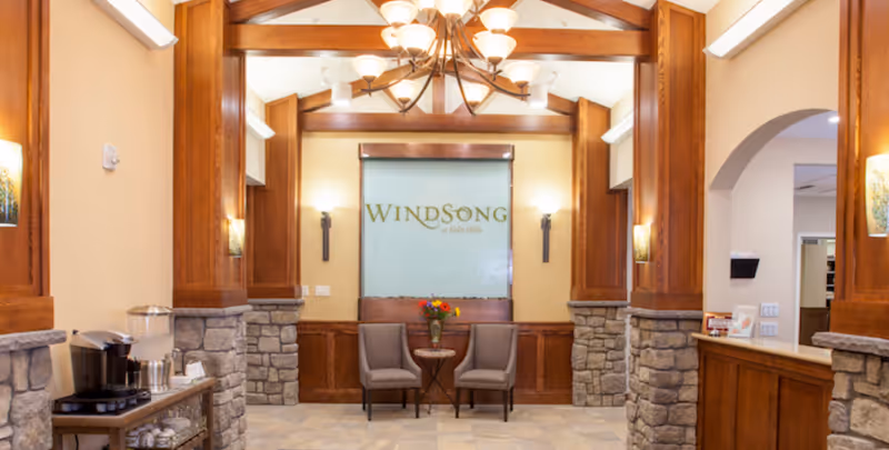 A cozy and elegant interior seating area with two gray chairs and a small round table with a vase of flowers between them. The background features a glass panel with the text 'Windsong at Eola Hills'. The room has warm lighting, wooden beams on the ceiling, stone pillars, and a chandelier hanging from the ceiling. To the left, there is a coffee station with a coffee maker and cups.