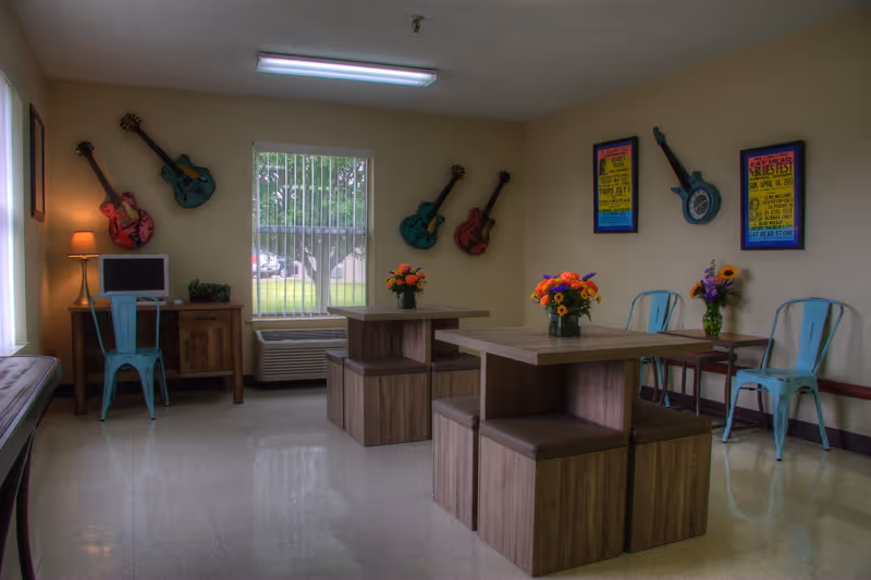 A bright room with two wooden tables and matching benches, each table decorated with a vase of colorful flowers. The walls are adorned with decorative guitars and framed posters. There are light blue metal chairs along the walls and a small desk with a computer and lamp near a window with vertical blinds. The floor is tiled and the room has a cozy, welcoming atmosphere.