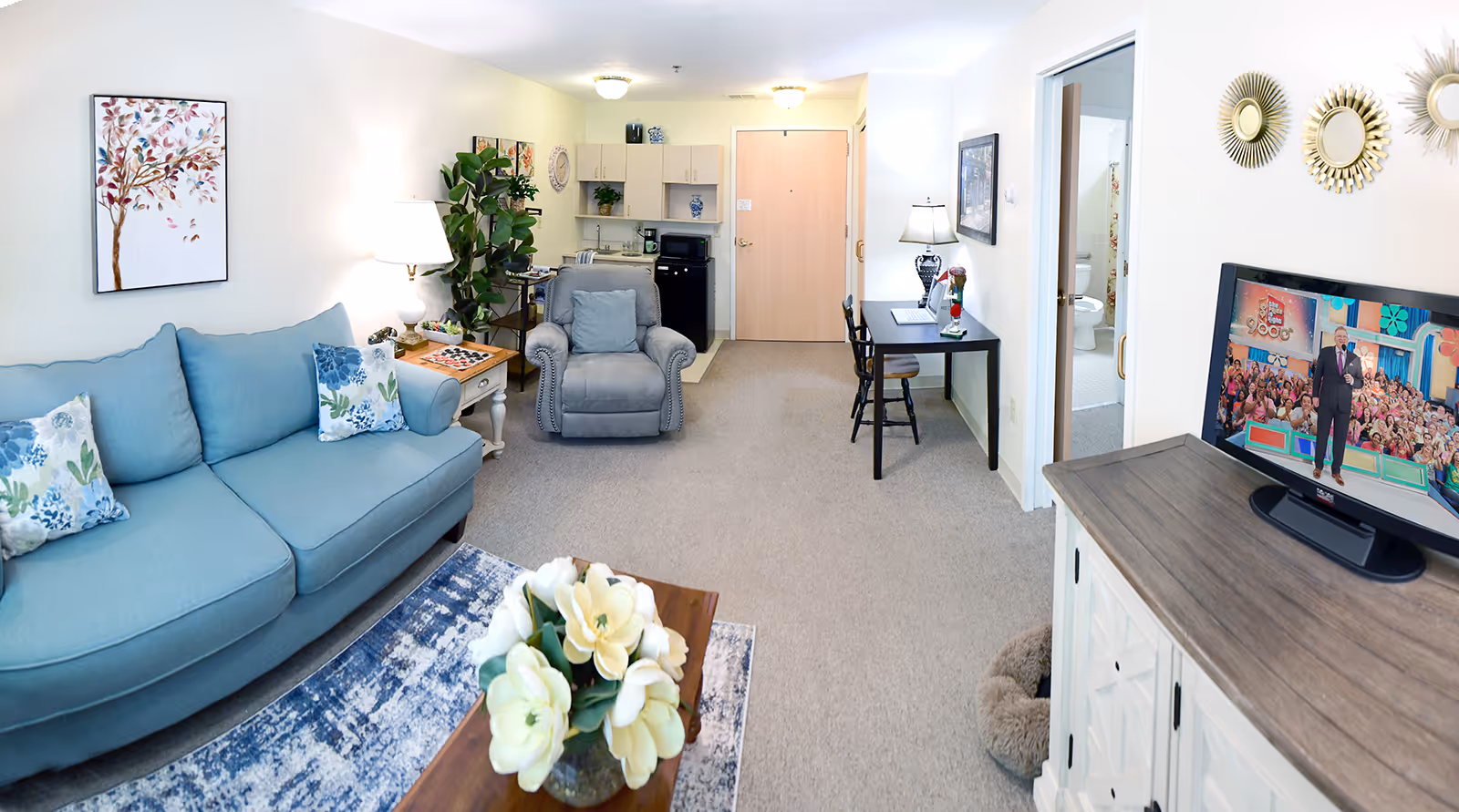 A cozy living room area in a senior living facility with a light blue sofa adorned with floral pillows, a gray armchair, a wooden coffee table with a vase of white flowers, and a TV on a white cabinet showing a game show. The room has beige carpet, a small desk with a chair and lamp, a kitchenette in the background, and a bathroom visible through an open door. The walls are decorated with artwork and mirrors.