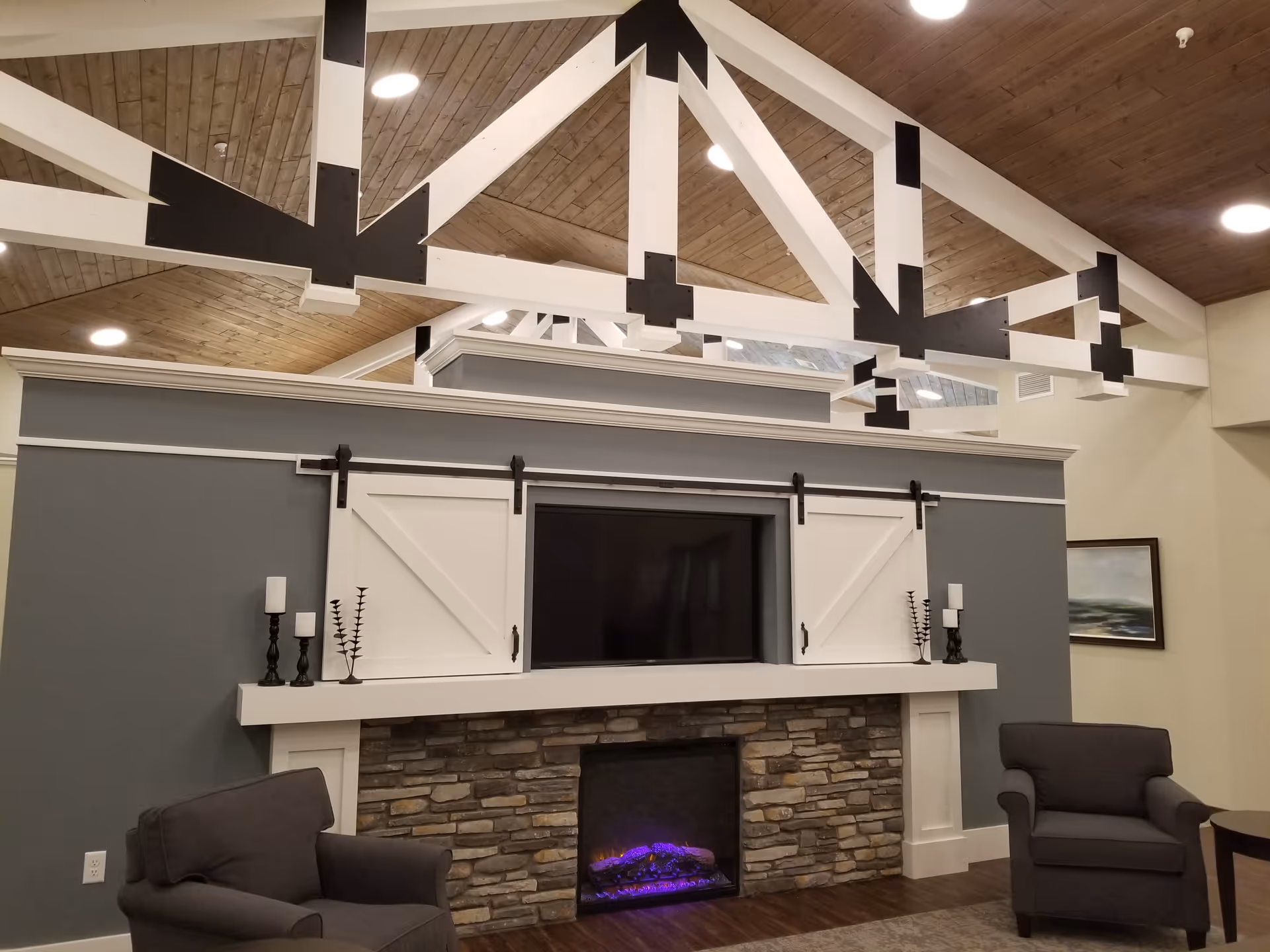 Cozy senior living common room with a stone fireplace and electric fire, TV framed by sliding barn doors, two armchairs, and exposed white ceiling beams.