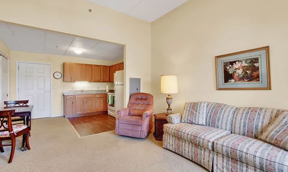 A cozy living area with a striped sofa, a pink recliner chair, a wooden side table with a lamp, and a framed floral painting on the wall. Adjacent to the living area is a small kitchen with wooden cabinets, a white refrigerator, a stove, and a sink. A dining table with chairs is partially visible on the left side.