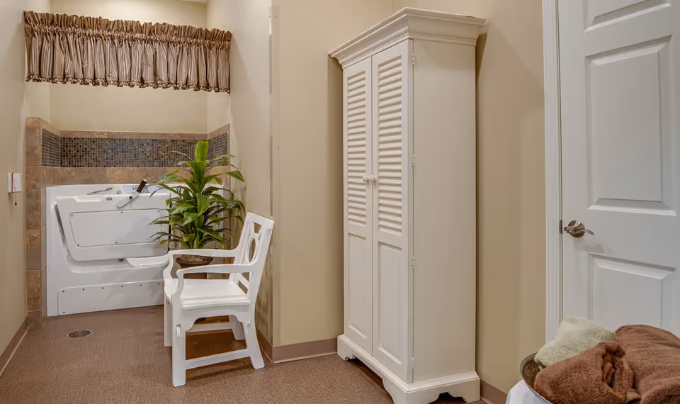 Accessible walk-in bathtub area with a white shower chair, potted plant, and a tall white cabinet in a beige room.