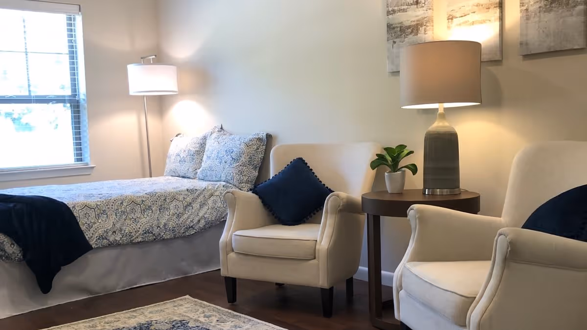 A cozy bedroom with a bed covered in patterned bedding and two matching pillows, a floor lamp beside the bed, two beige armchairs with dark blue cushions, a round wooden side table with a small potted plant and a table lamp, and a window with blinds letting in natural light.