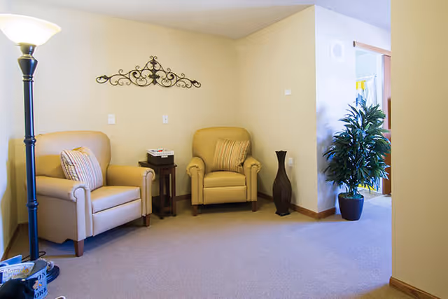 A cozy sitting area with two beige armchairs, each with a striped pillow, separated by a small wooden side table holding a tissue box. A tall floor lamp is on the left side, and a decorative metal wall art piece is mounted on the light yellow wall behind the chairs. To the right, there is a large potted plant near an open doorway leading to another room.
