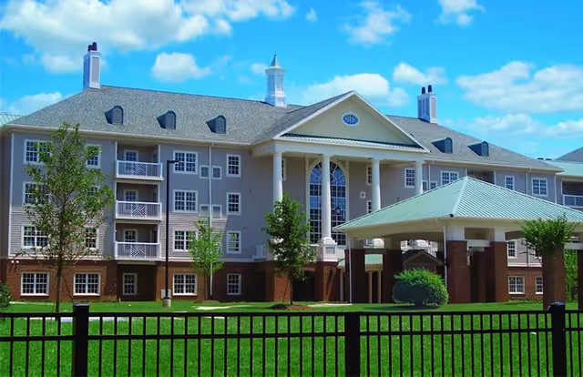 Exterior view of a large senior living facility building with multiple windows, balconies, and a covered entrance. The building has a gray roof with chimneys and is surrounded by a green lawn and a black metal fence. The sky is blue with scattered clouds.
