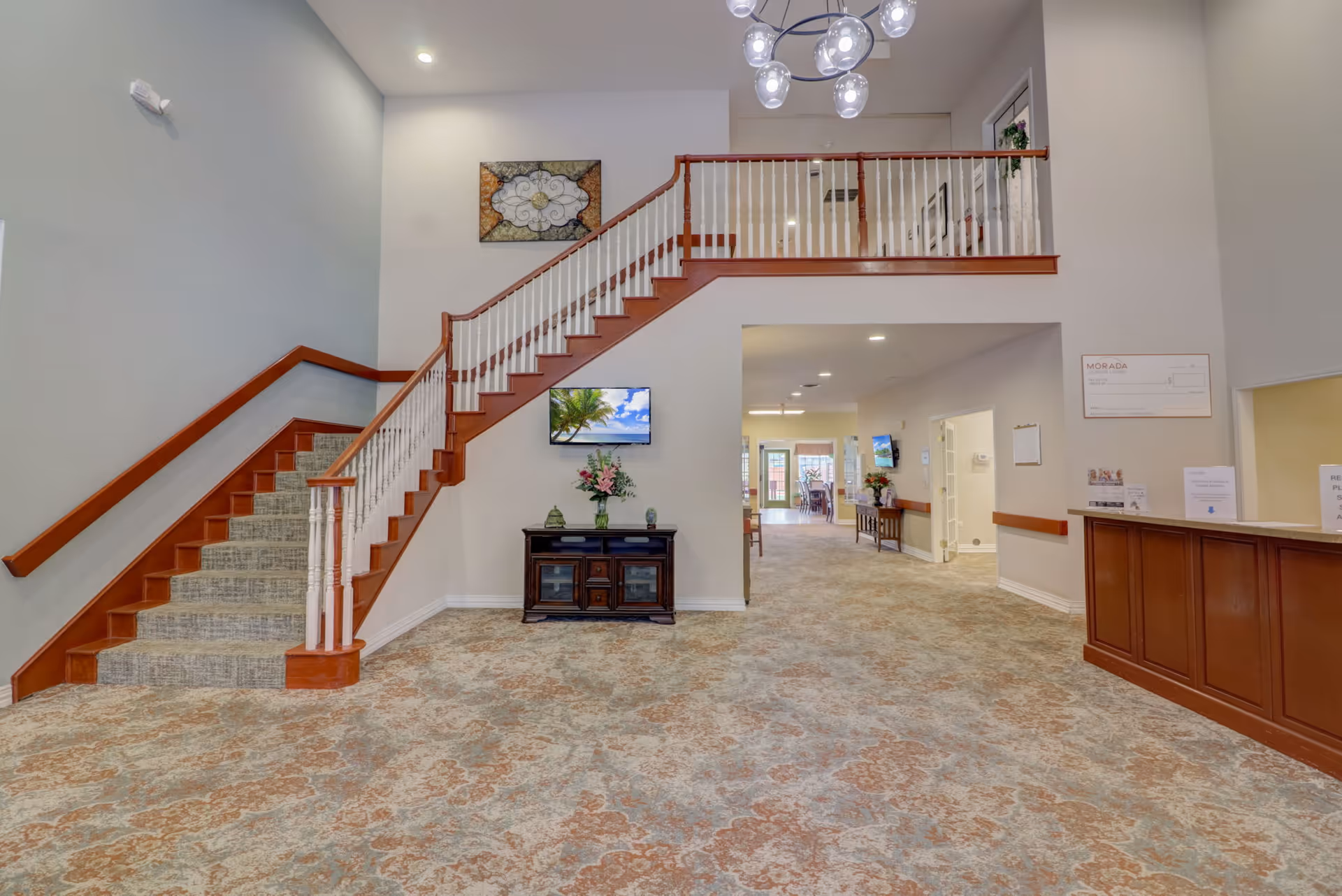 Interior view of a senior living facility lobby area with a carpeted floor, a wooden staircase with white balusters leading to an upper floor, a small dark wooden cabinet with a vase of flowers and decorative items, a wall-mounted TV displaying a beach scene, and a reception desk on the right side. The hallway beyond the lobby leads to a dining area with tables and chairs visible in the distance.