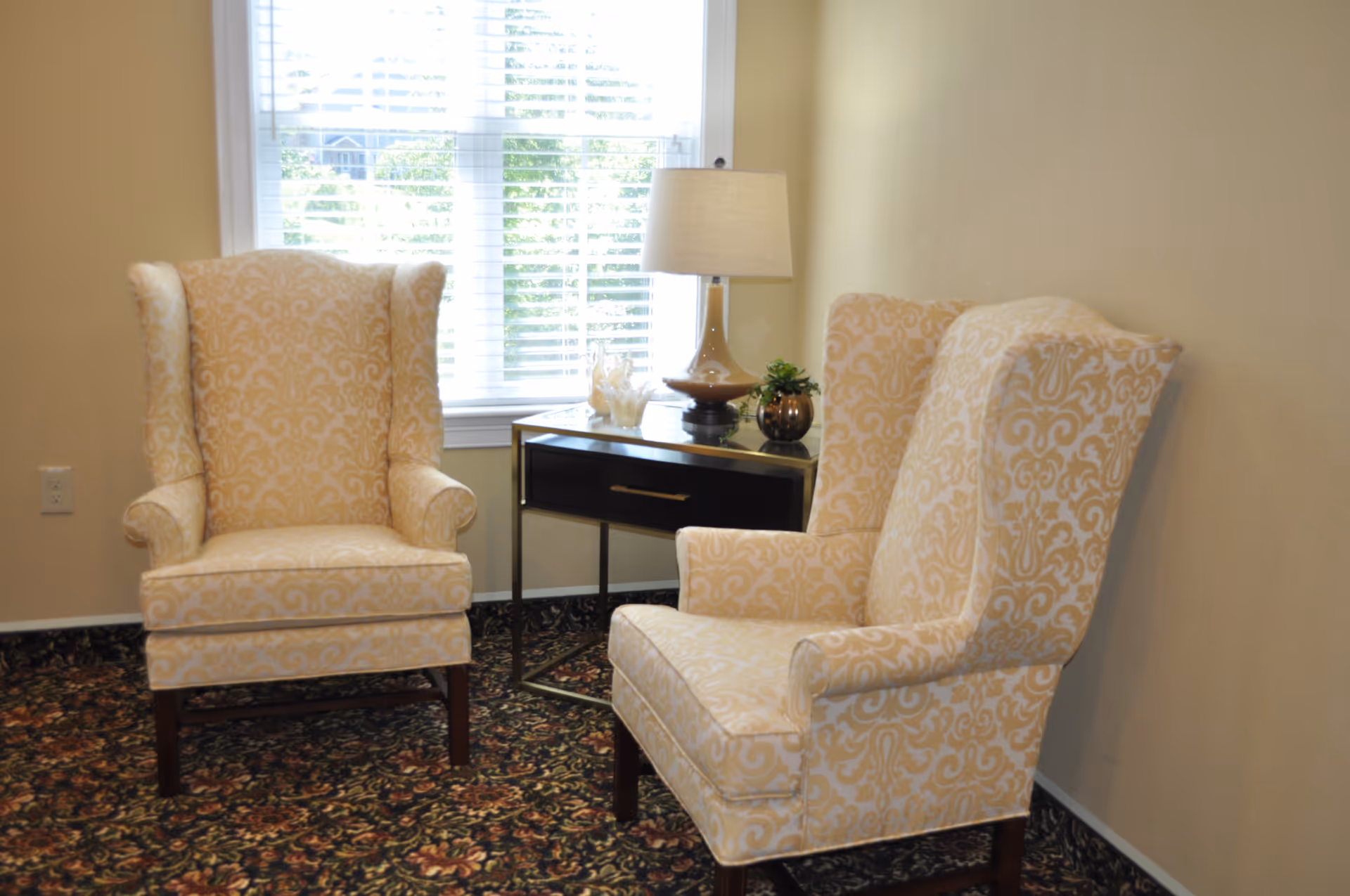 Two patterned wingback chairs flank a small side table with a lamp in a bright sitting area beside a window.