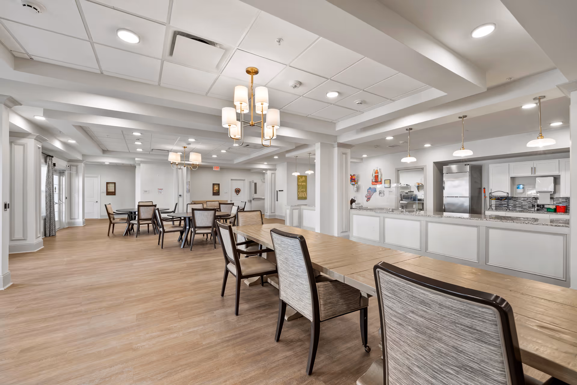A spacious, well-lit dining area in a senior living facility with multiple tables and chairs arranged neatly. The room features light wood flooring, white walls, and a ceiling with recessed lighting and chandeliers. On the right side, there is a kitchen counter with pendant lights hanging above, stainless steel appliances, and a serving area.