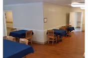 Interior view of a dining area in an assisted living facility with several wooden tables covered with blue tablecloths and wooden chairs arranged around them. The room has light-colored walls and wood flooring, with doorways visible in the background.