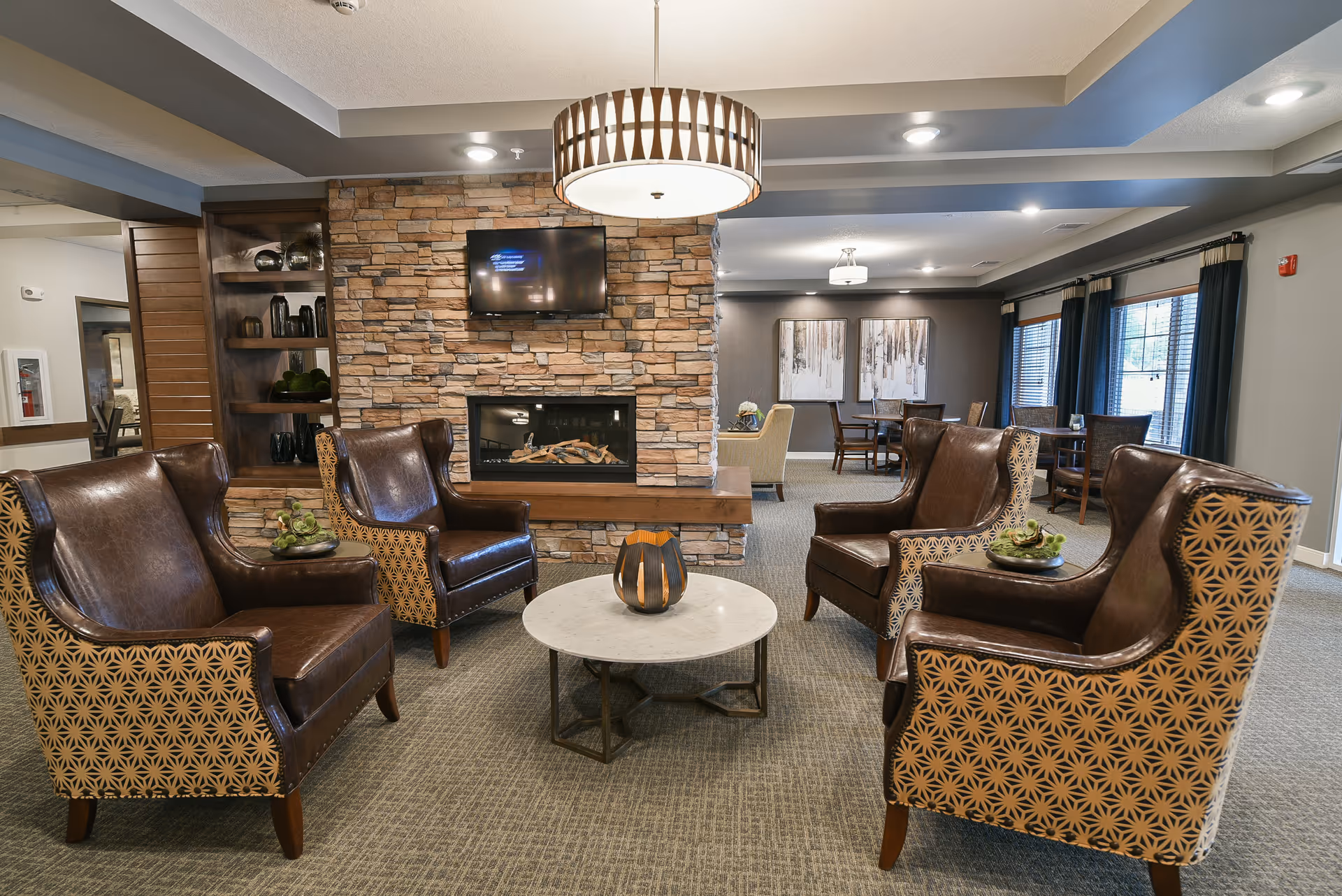 A cozy lounge with four leather armchairs around a round coffee table facing a stone fireplace and wall-mounted TV.