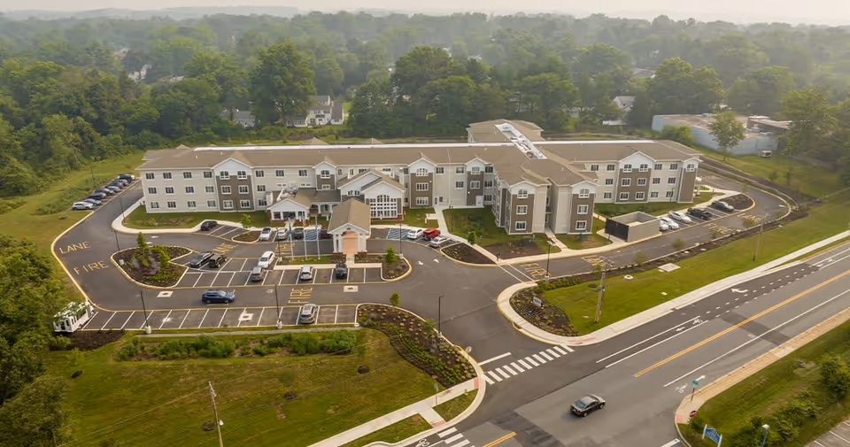Aerial view of Juniper Village at Newark, showing a large three-story senior living facility surrounded by parking lots, landscaped areas, and roads. The building is beige and white with multiple windows and entrances. The surrounding area includes green trees and a road with marked lanes and crosswalks.