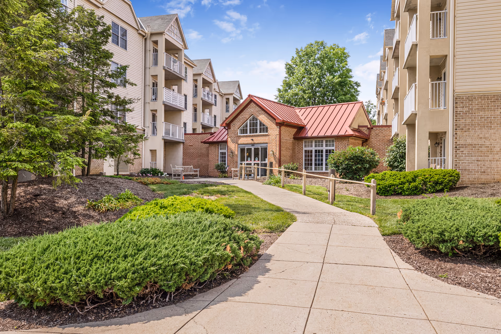 Outdoor view of a senior living facility with a paved walkway leading to a small brick building with a red roof. The building is surrounded by green bushes, trees, and landscaped garden beds. On either side of the walkway are multi-story residential buildings with balconies. The sky is blue with some clouds.