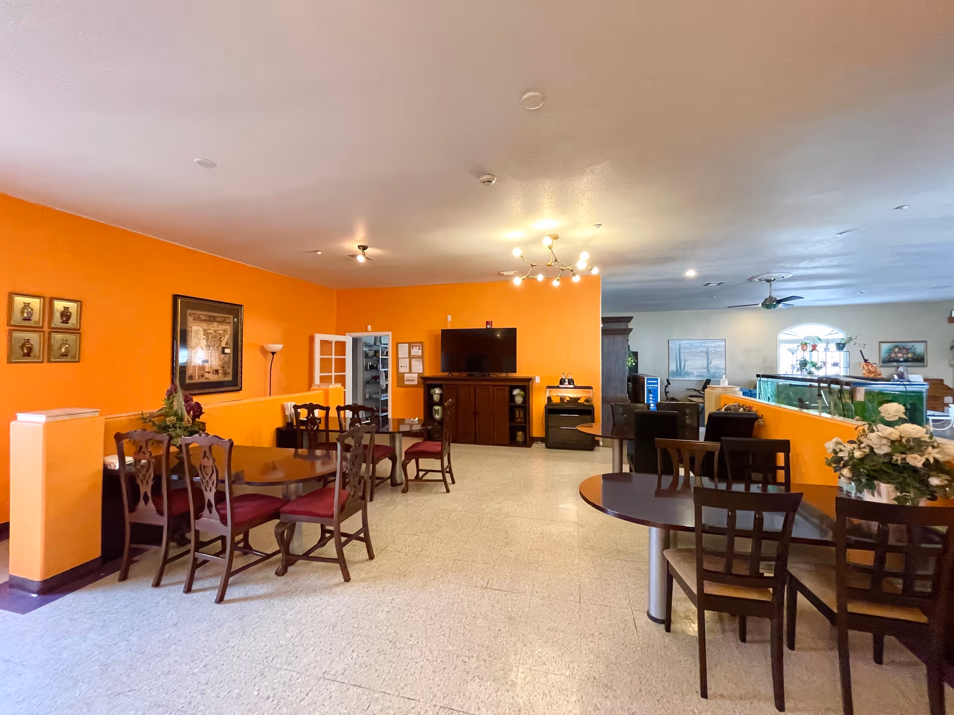 Bright communal dining area with multiple wooden tables and chairs, orange walls, and a TV on a cabinet.