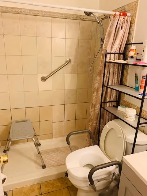 A bathroom with a tiled shower area featuring a grab bar and a shower chair. There is a toilet with armrests and a shelving unit above it holding various toiletries. A shower curtain is partially drawn to the side.