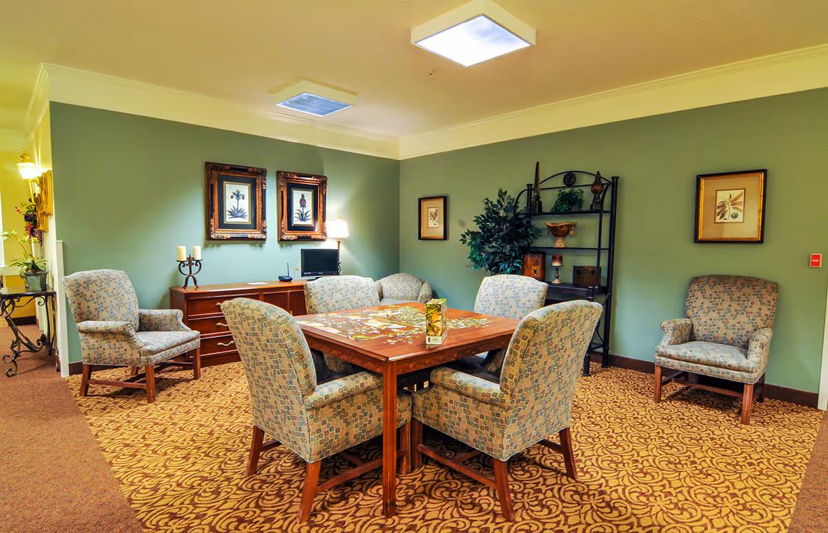 Communal seating area with a square wooden table surrounded by upholstered chairs in a softly lit room with green walls and framed artwork.