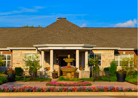 Front exterior view of a single-story brick building with a covered entrance supported by columns, a central fountain, benches, potted plants, and colorful flower beds under a clear blue sky.