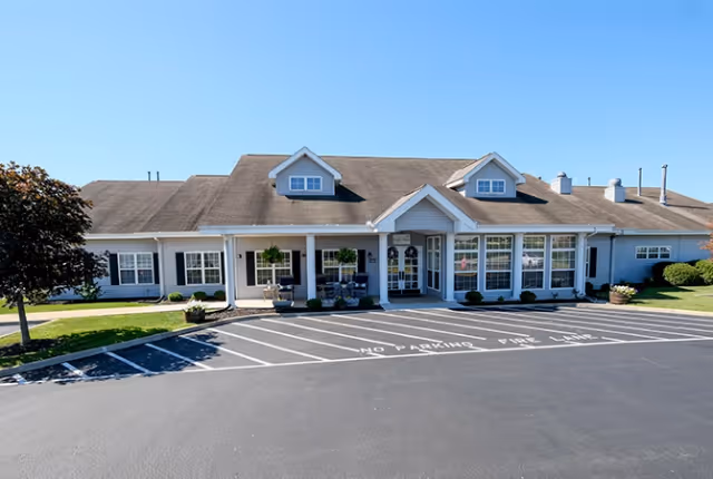 Front exterior view of a single-story building with a pitched roof, multiple windows, and a covered entrance. There is a parking lot in front with marked no parking zones and a tree on the left side of the building. The sky is clear and blue.