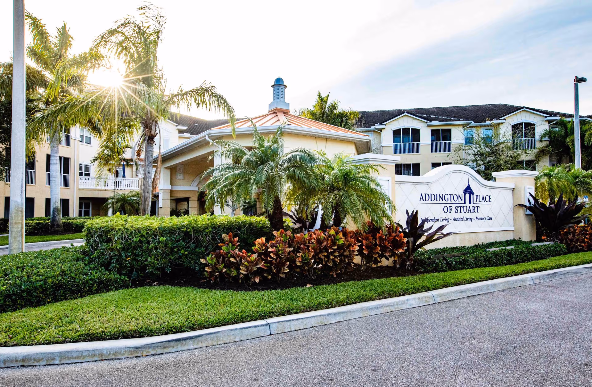 Exterior view of Addington Place of Stuart senior living facility with a landscaped entrance featuring palm trees, bushes, and colorful plants. The building is a multi-story structure with balconies and a covered entrance. The sun is shining through the palm trees.