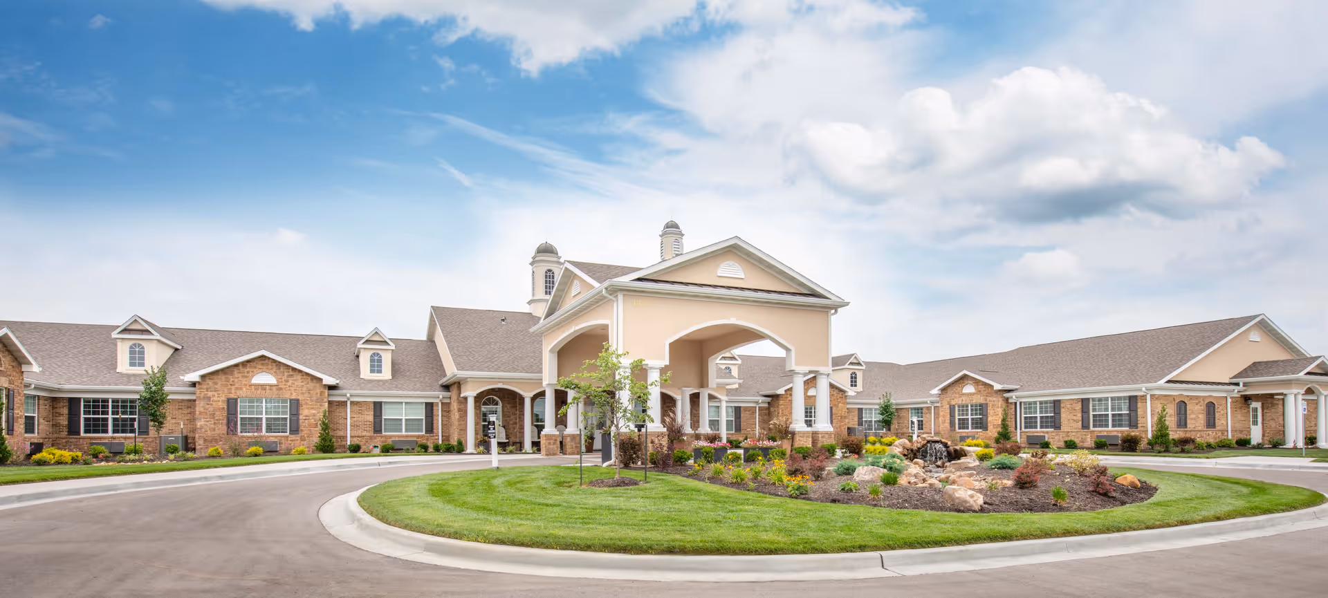 Front exterior of a single-story senior living building with a covered porte-cochere, circular driveway, and landscaped lawn.