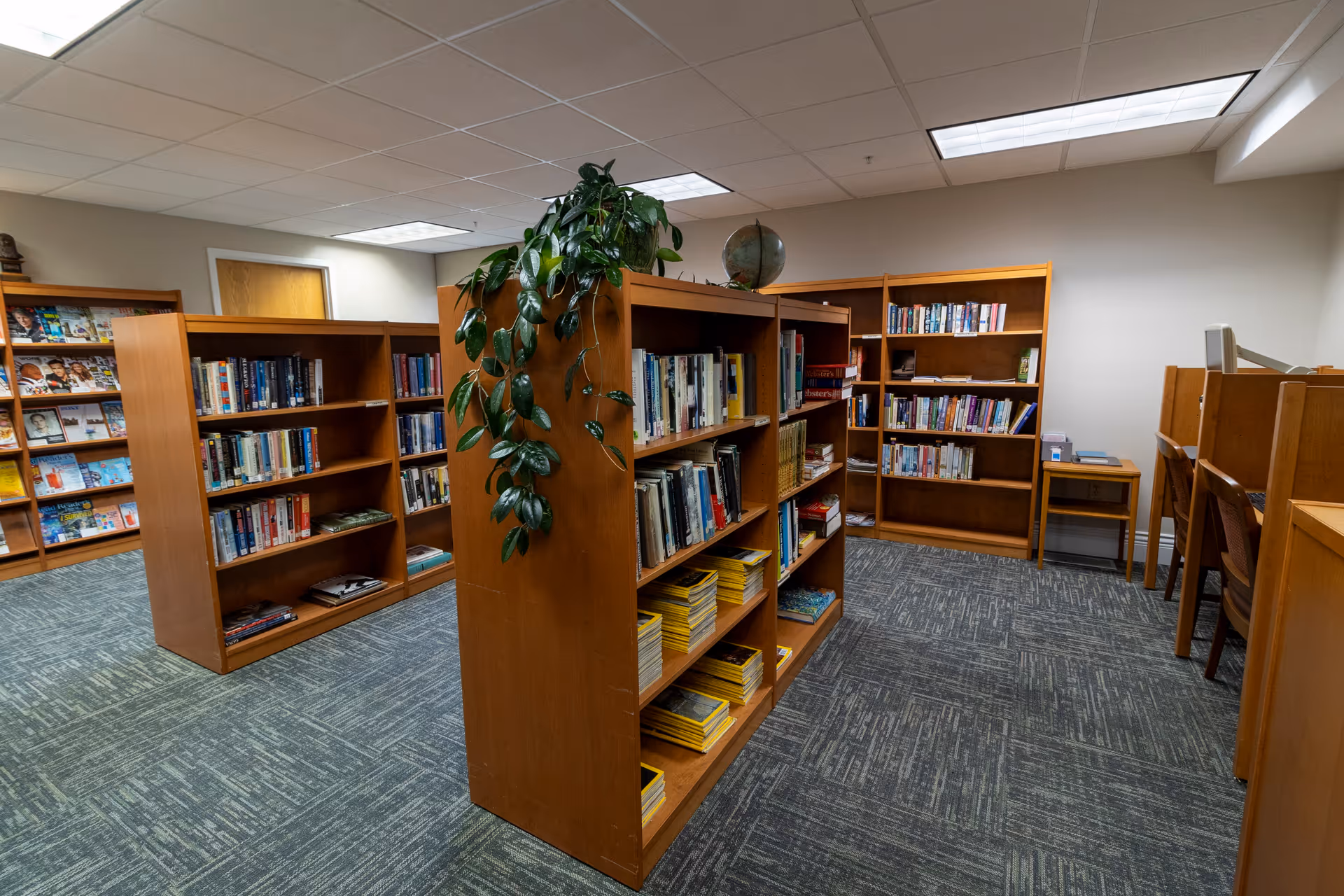 Interior view of a library or reading room with wooden bookshelves filled with books and magazines. There is a green leafy plant on top of one bookshelf and a globe. On the right side, there are wooden study carrels with chairs. The room has a carpeted floor and a drop ceiling with fluorescent lights.