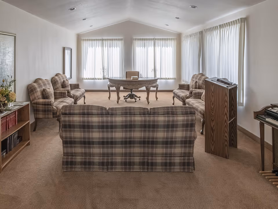 A spacious living room with beige carpet and white walls, featuring plaid upholstered sofas and armchairs arranged around a wooden desk with a swivel chair near large windows covered with sheer white curtains. There is a wooden bookshelf with books and a flower arrangement on the left, and a wooden podium and electronic keyboard on the right.