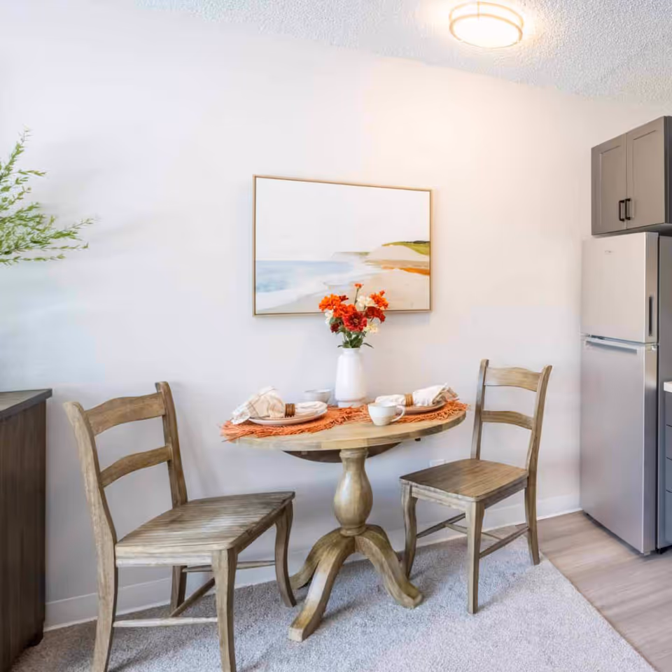 Small dining nook with a round wooden table set for two, two wooden chairs, a vase of red flowers, wall art, and a nearby refrigerator.