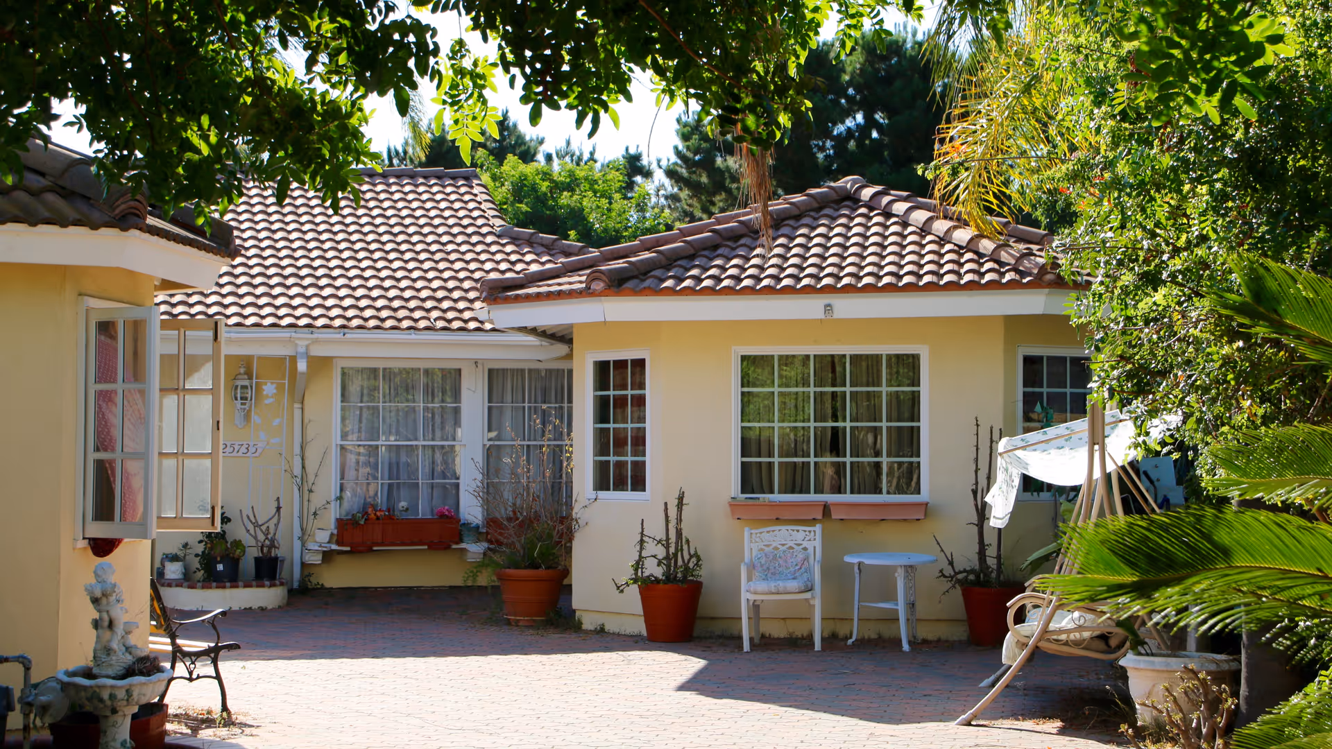 Yellow single-story house with a tiled roof and a sunlit brick courtyard featuring potted plants, chairs, and a small fountain.