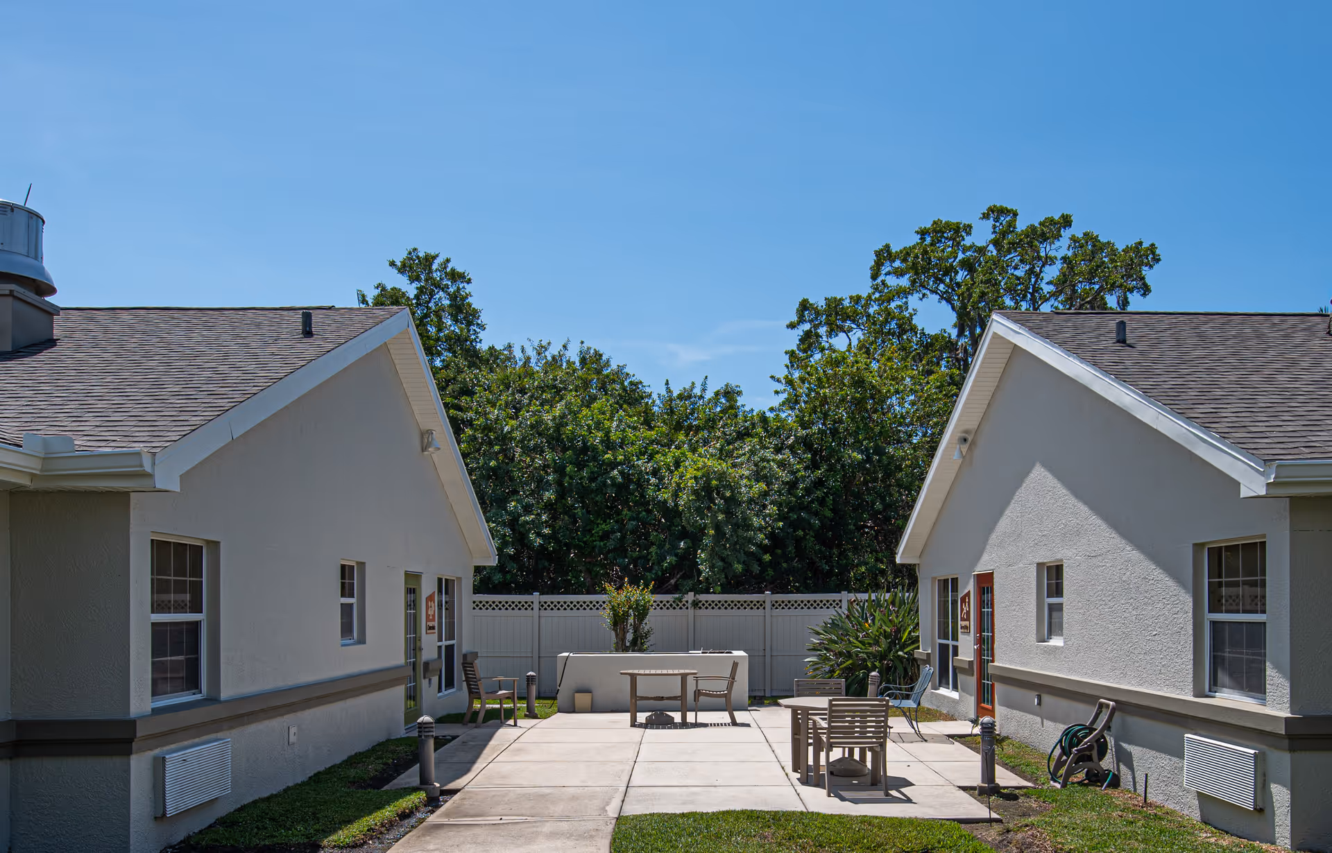 Outdoor courtyard area between two single-story buildings with beige walls and brown shingle roofs. The courtyard has a concrete walkway and patio with several wooden chairs and tables. There is a white fence and green trees in the background under a clear blue sky.