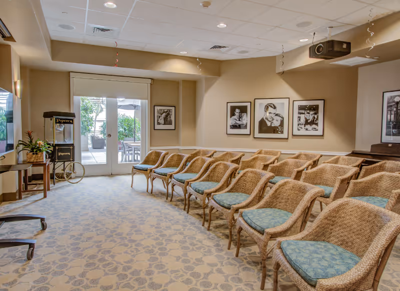 Community activity room with rows of wicker chairs facing a screen, framed black-and-white photos on the wall, a popcorn cart, and double doors to an outdoor patio.