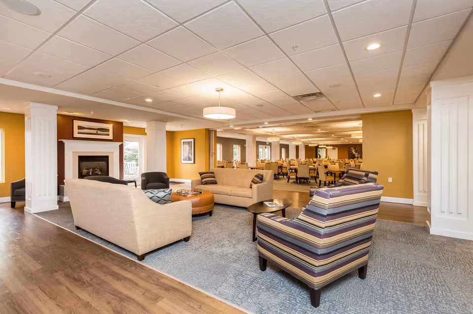 A spacious senior living facility common area with beige sofas, a striped armchair, a round ottoman, and a fireplace. The room has warm yellow walls, wooden flooring, and a carpeted seating area. In the background, there is a dining area with multiple tables and chairs.