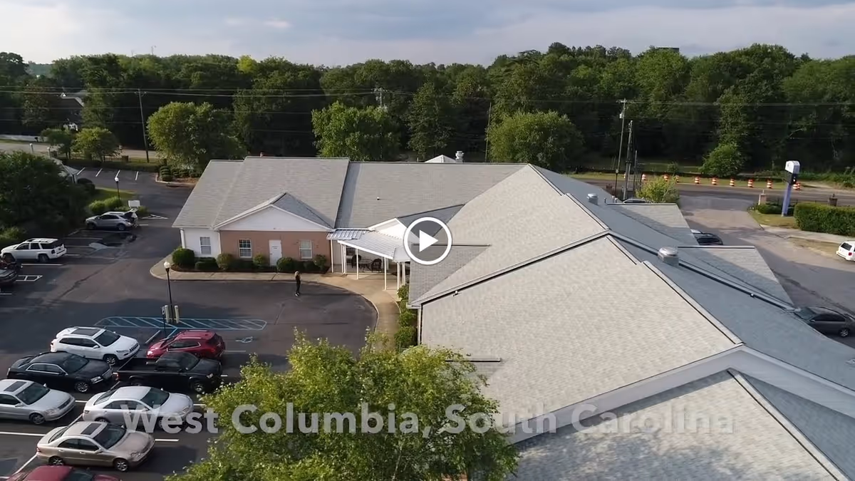 Aerial view of a single-story building with a gray roof and a parking lot with several cars parked. The building is surrounded by trees and greenery, with a road visible in the background. The text 'West Columbia, South Carolina' is overlaid on the image.