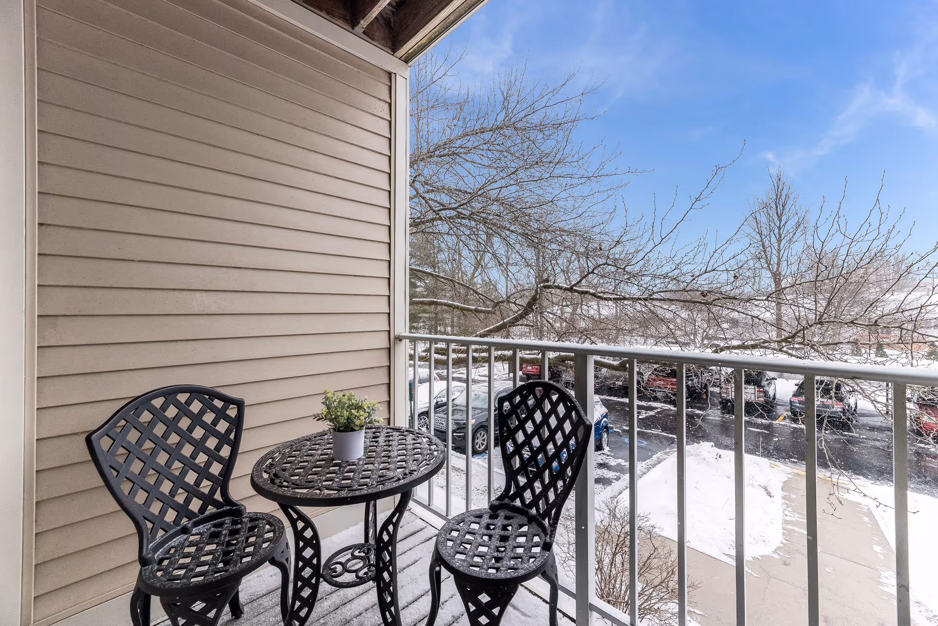 A small outdoor balcony with a black metal table and two matching chairs. A small potted plant is placed on the table. The balcony overlooks a parking lot with several cars and leafless trees, with patches of snow on the ground and a clear blue sky above.