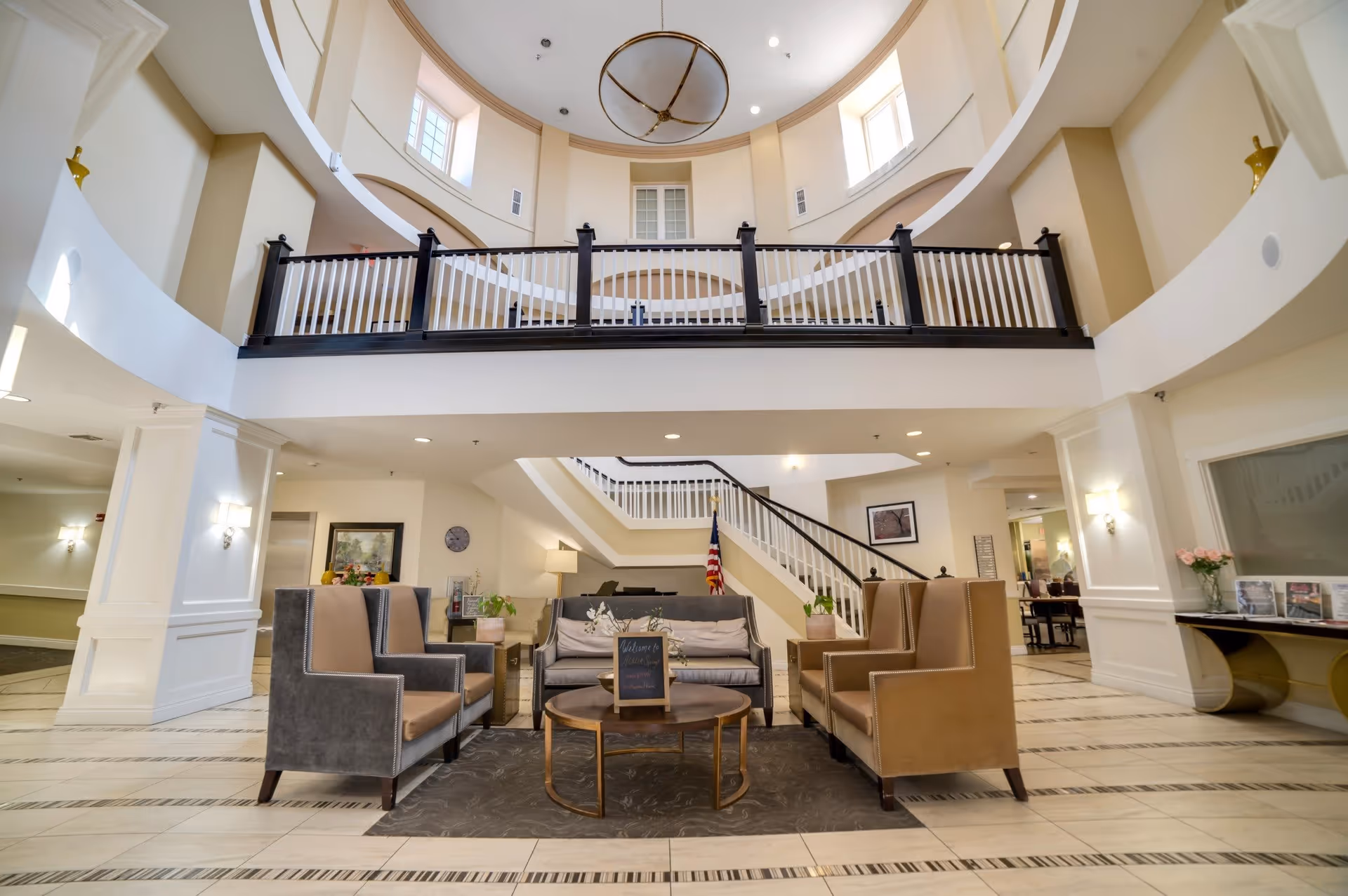 Spacious and well-lit lobby area with a high ceiling and a large circular chandelier. The seating area includes a gray sofa, two gray armchairs, and two tan armchairs arranged around a round coffee table with a small chalkboard sign. A staircase with white railings and dark handrails leads to the upper floor. The walls are painted white with decorative molding, and there are windows near the ceiling allowing natural light to enter.