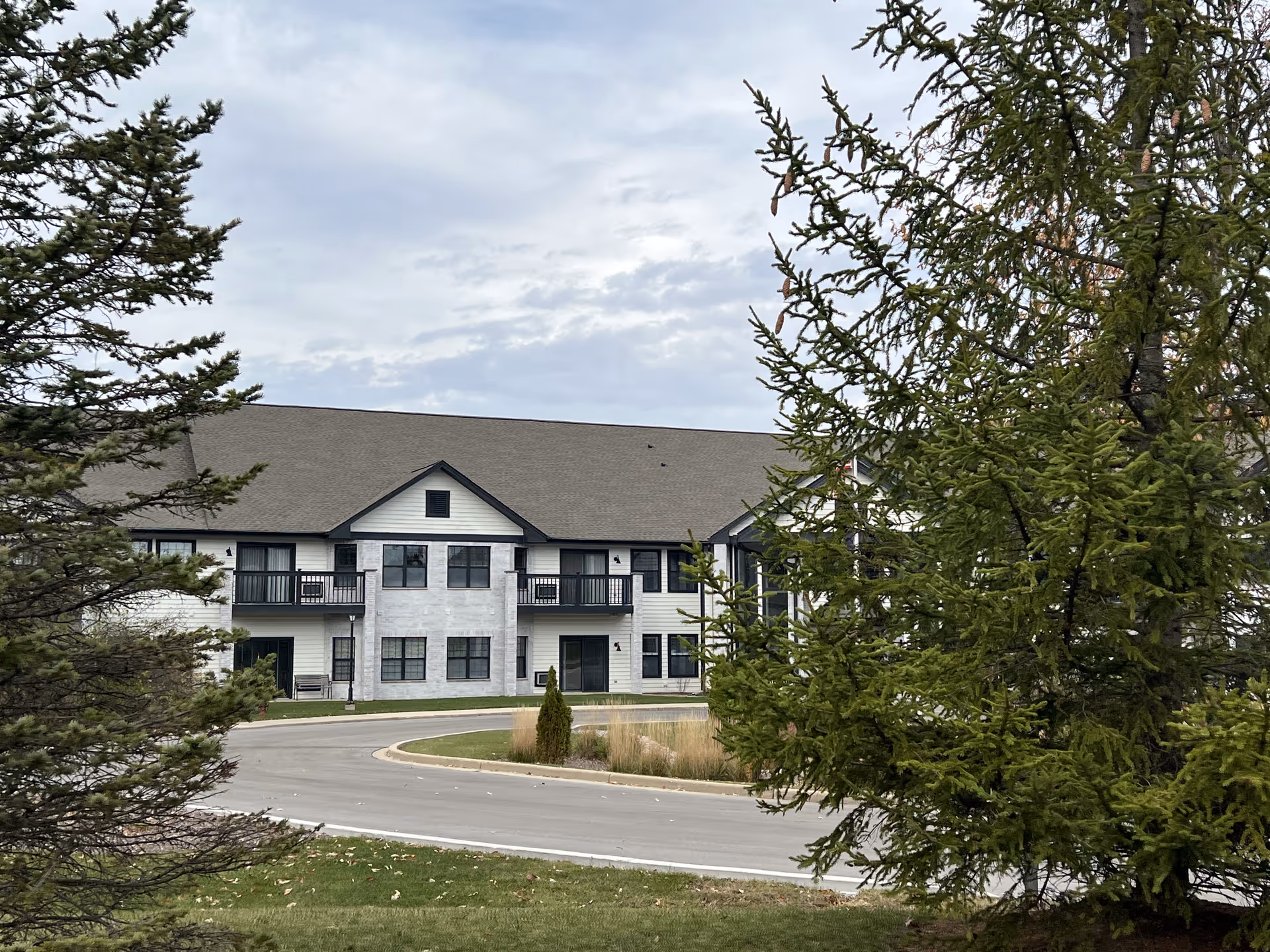 Exterior view of a two-story senior living facility building with a sloped roof, multiple windows, and balconies. The building is partially obscured by evergreen trees in the foreground, with a curved driveway and landscaped grass area in front.