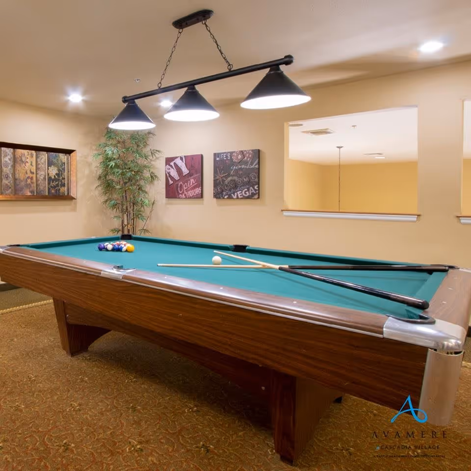 A recreational room with a pool table featuring a green felt surface, pool balls arranged in a triangle, and two pool cues resting on the table. Above the table, there is a black hanging light fixture with three lamps. The room has beige walls decorated with framed artwork and a tall green plant in the corner. There is a large rectangular opening in the wall providing a view into another room.