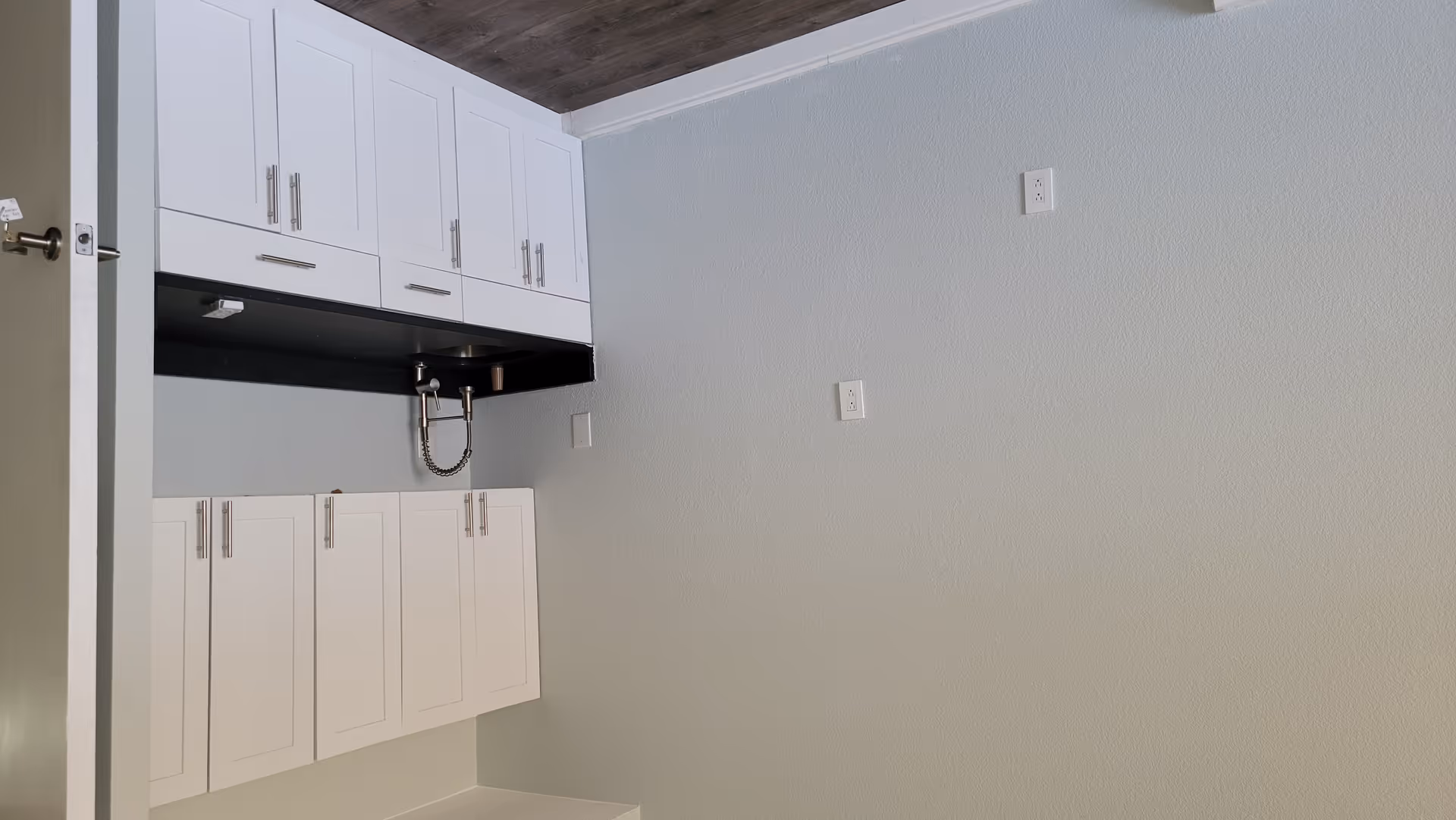 A small kitchenette area with white cabinets, a black countertop, and a modern faucet with a flexible spring neck. The walls are painted light gray, and there are multiple electrical outlets on the wall. The floor has a wood-like finish, and a partially open door is visible on the right side.