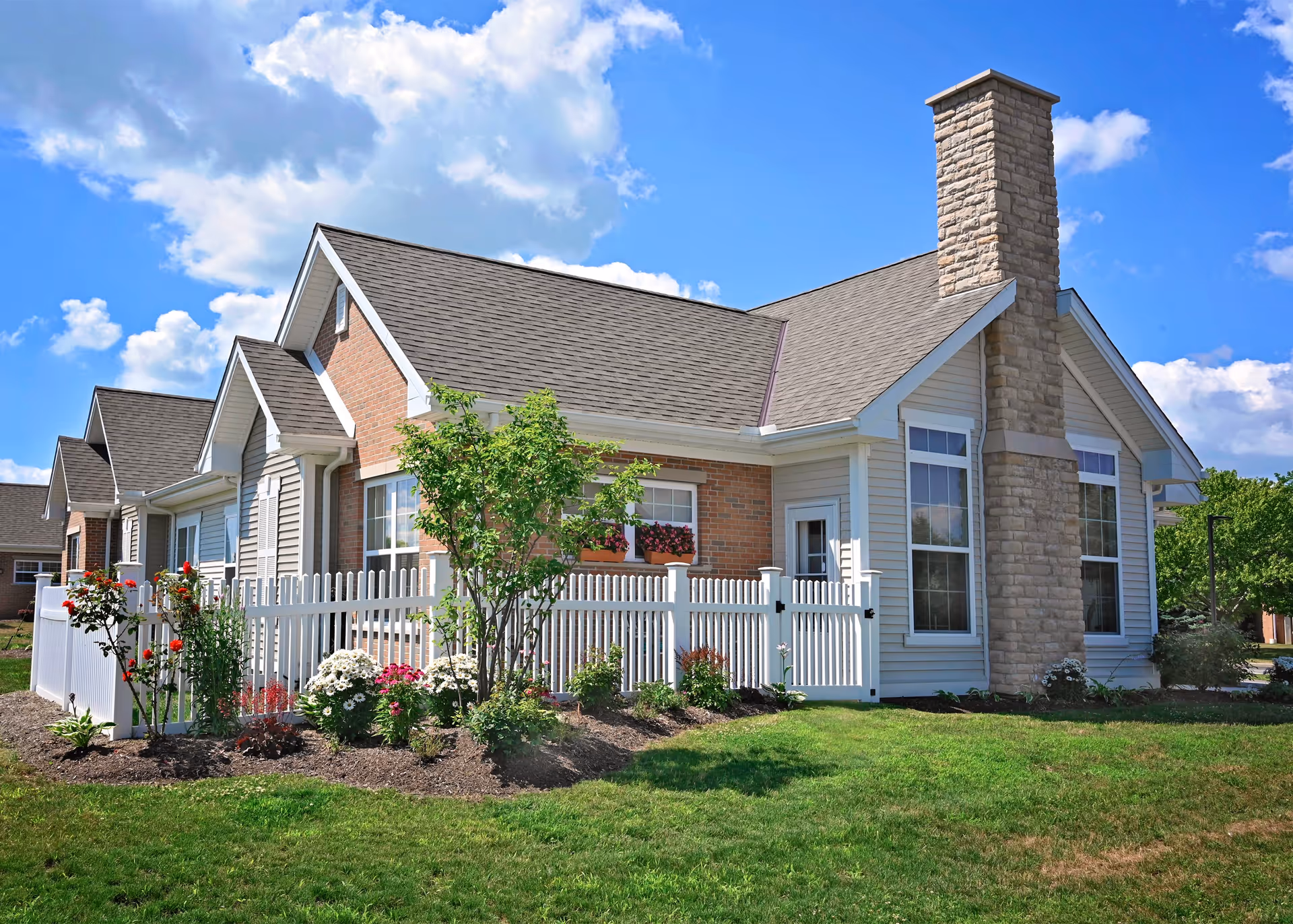 A single-story residential building with a brick and siding exterior, a stone chimney, and multiple gabled roofs. The building is surrounded by a white picket fence enclosing a garden with various flowers and shrubs. The sky is blue with scattered clouds.