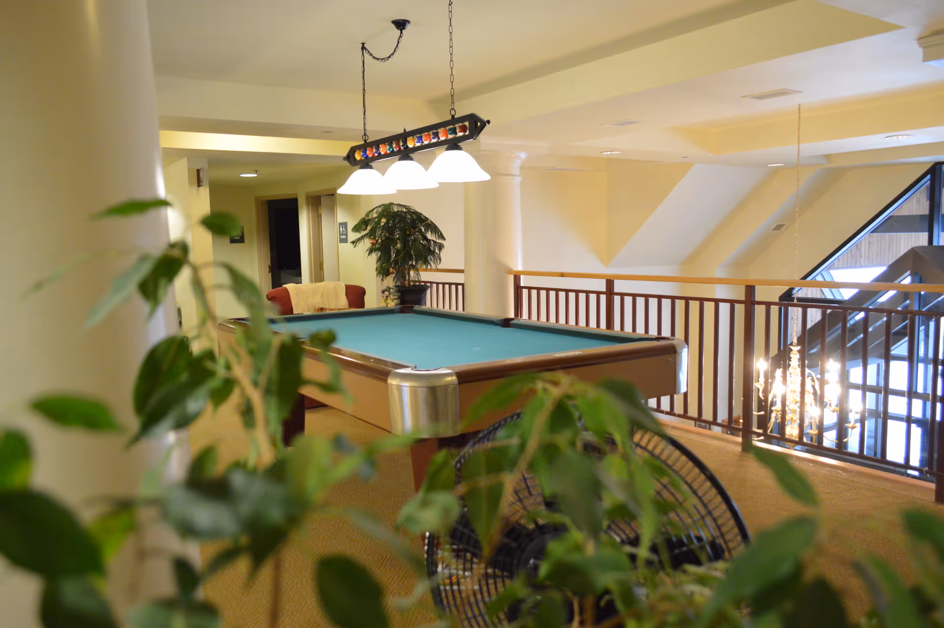 Interior view of a recreational area in a senior living facility featuring a pool table with a hanging light fixture above it. There are plants in the foreground and background, a red armchair with a blanket, and a railing overlooking a lower level with a chandelier visible.
