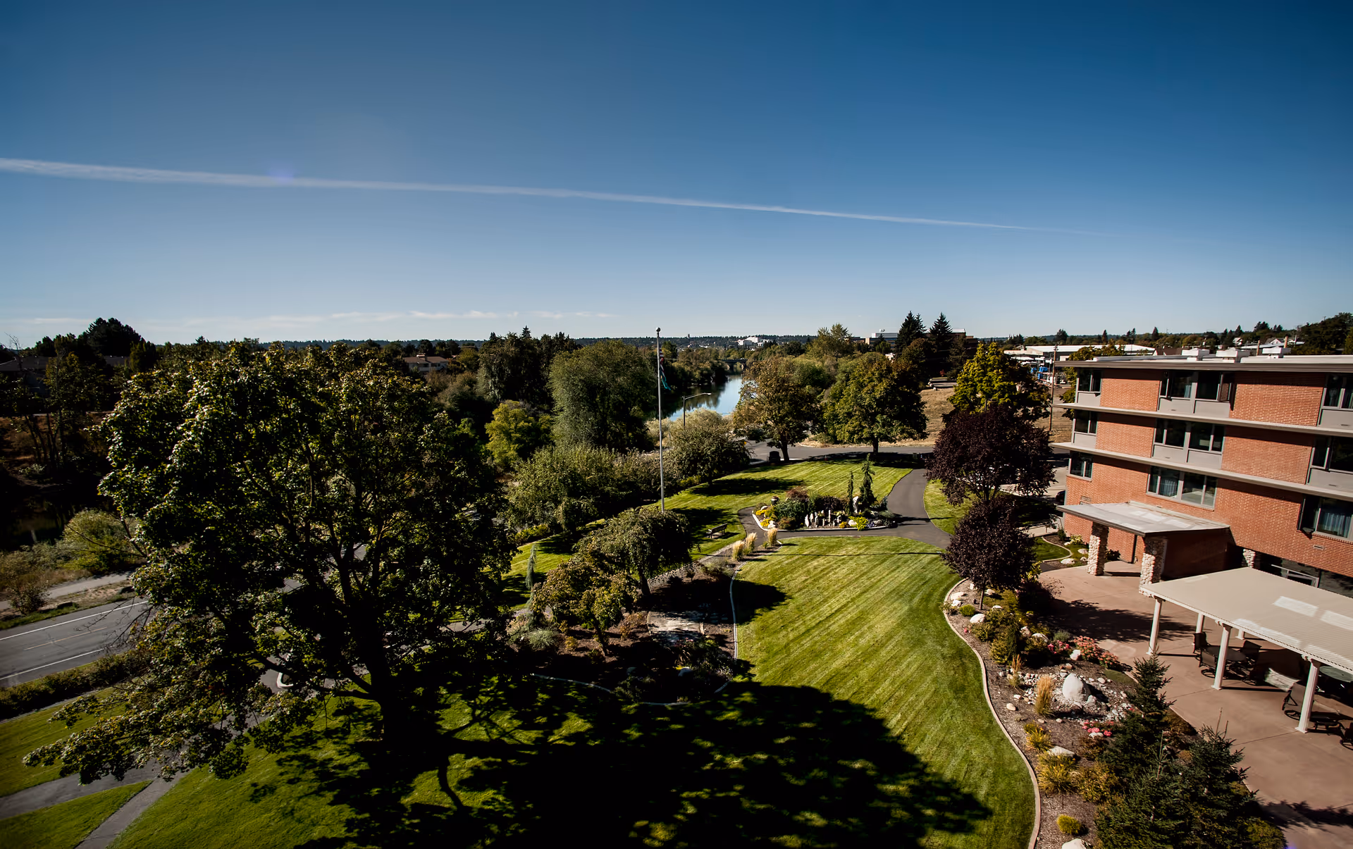 Aerial view of Riverview Terrace senior living facility showing a large green lawn with trees, landscaped garden beds, a flagpole, paved walkways, and a multi-story brick building on the right. In the background, a river runs through a wooded area under a clear blue sky.