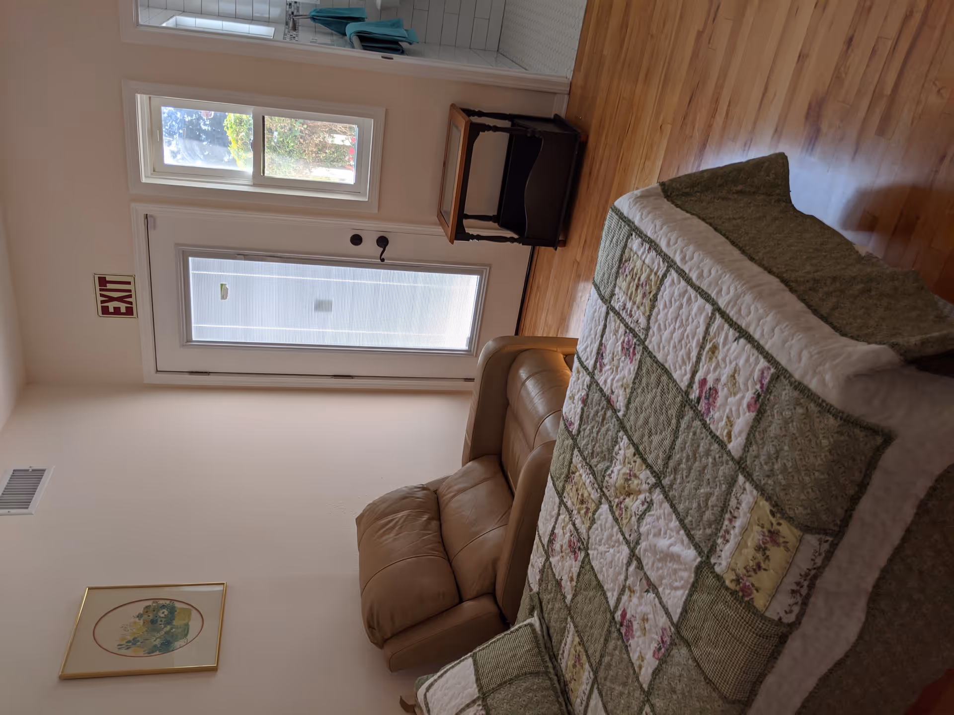 A cozy room with a bed covered in a green and white patchwork quilt, a beige leather armchair, a small wooden side table, a window, and a door with an exit sign above it. The floor is wooden, and there is a framed picture on the wall.