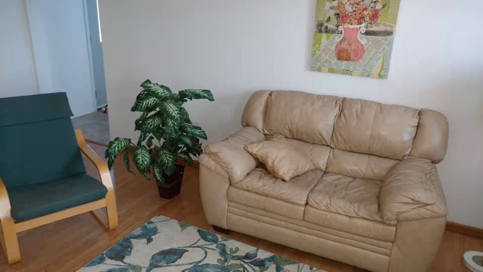 A cozy living room area featuring a beige leather loveseat with a matching pillow, a green cushioned wooden armchair, a potted green plant, a floral patterned rug on a wooden floor, and a colorful floral painting hanging on a white wall.
