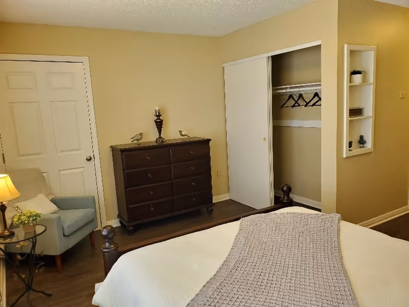 A bedroom with a bed covered in a white blanket and a gray knitted throw. There is a dark wooden dresser with decorative items on top, a light blue armchair with a pillow, a small round side table with a lamp and a plant, a closet with sliding white doors partially open showing hangers, and a built-in wall shelf with small decorative items. The walls are painted beige and the floor is wooden.