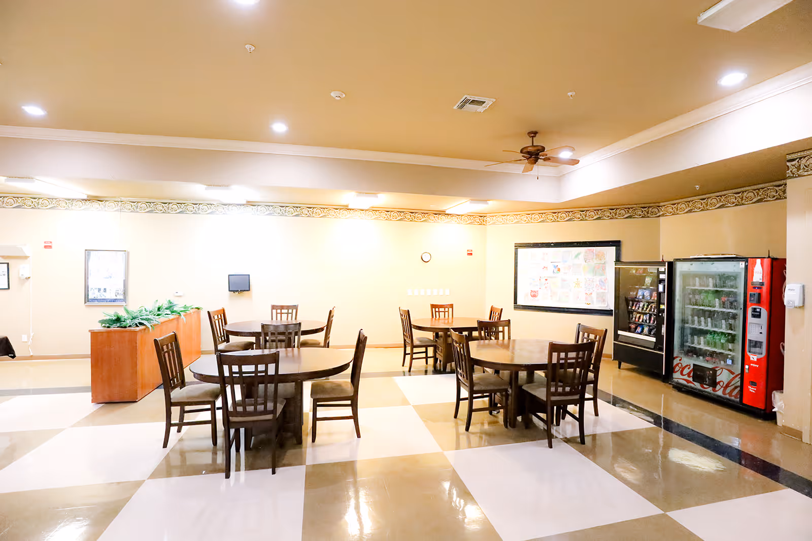 A bright and clean common dining area with several round wooden tables and chairs arranged neatly. There are vending machines against the wall on the right side and a bulletin board with colorful papers displayed. The floor has a checkered pattern with beige and white tiles, and the ceiling has recessed lighting and a ceiling fan.